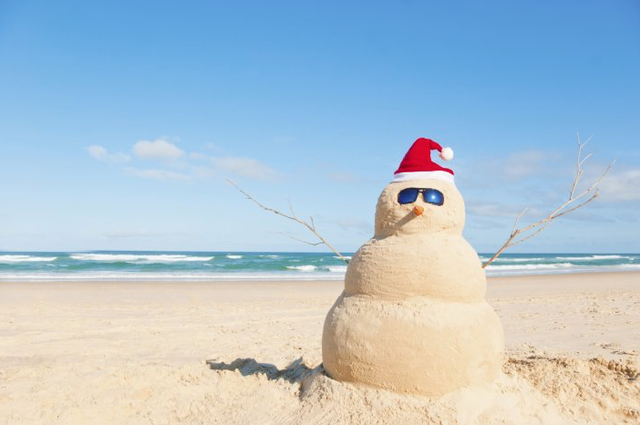 A photo of a snowman made out of sand on a beach, with blue skies overhead. The Snowman is wearing a red hat and sunglasses.